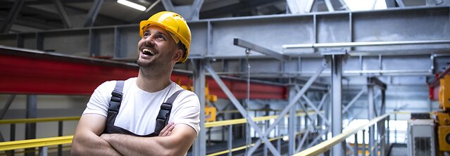 Portrait of smiling factory worker with arms crossed standing in industrial production hall.